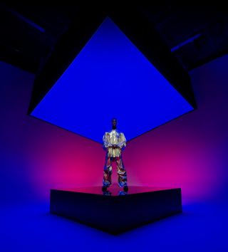 A person dressed in a metallic suit standing on a square centre stage and encompassing the background showcasing the Ultra Wide camera on iPhone 16 Pro Max